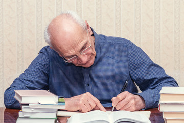 an elderly male scientist makes notes in a notebook, books on the table