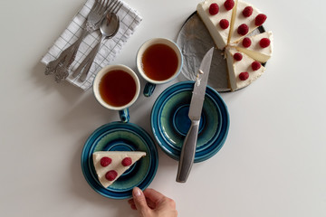 View from above on woman hands taking plate with raspberry cheesecake piece. Sliced cake and cups with tea on served white table background