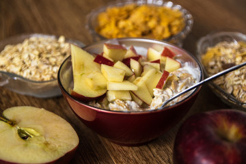 Muesli with dried fruit, milk and sliced red apple on wooden table. Barley flake, fresh organic red apple,  and corn flakes in the background.