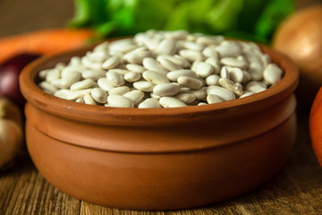 Uncooked white bean in a clay bowl on wooden table. Tomato, red onion, garlic, carrot and lettuce in the background.