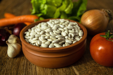 Uncooked white bean in a clay bowl on wooden table. Tomato, red onion, garlic, carrot and lettuce in the background.