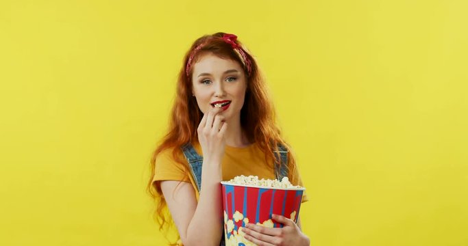 Beautiful young woman with long red hair holding a big popcorn and eating it in front of the camera on the yellow wall background.