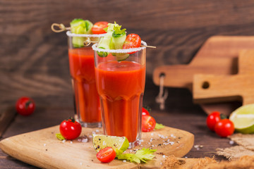 Two glasses of tomato juice decorated with fresh tomatoes, cucumber and leaves on a wooden background
