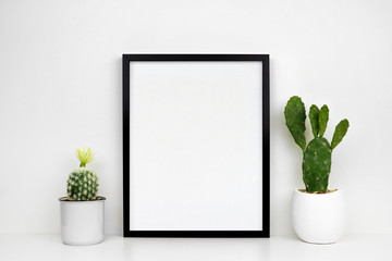 Mock up black frame with cactus plants in pots on a shelf or desk. White shelf and wall. Portrait frame orientation.