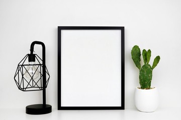 Fototapeta premium Mock up black frame, cactus plant and industrial style lamp on a shelf or desk. White shelf and wall. Portrait frame orientation.