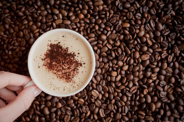  female hand with a neat natural manicure holding a white cup of hot cappuccino on the background of coffee beans with free space on the right