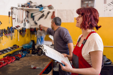 Cute, female mechanic in red work wear overalls writes on clipboard in car garage. Male mechanic and car mechanic tools in the background. Gender equality, diversity concept;