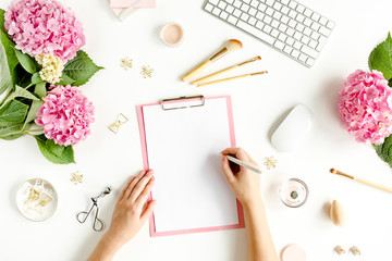 Female workspace with female hands, clipboard, bouquet hydrangea, computer, accessories on white background. Stylized women's desk. Flat lay. Top view.