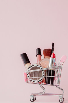 Creative concept with shopping trolley with makeup on a pink background. Perfume, sponge, brush, mascara, pencil, nail file, eye shadow, lip gloss in the basket, copy space