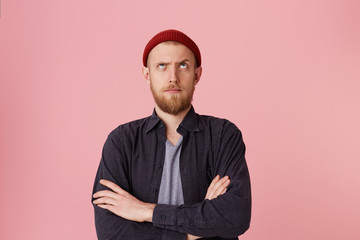Photo of questioning bearded man in basic shirt, with red hat, with folded arms, look up with one...