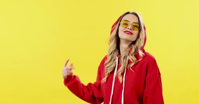 Pretty caucasian young stylish woman in red hoodie and sunglasses walking on the yellow background into the shooting frame and going away, then handsome guy doing the same.