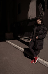 Fototapeta na wymiar Fashion beautiful man wearing in black jacket and trousers, red sneakers, backpack, glasses. Street city style