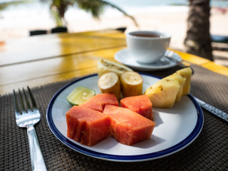Healthy fruit breakfast on sunny beach. Cup of tea. Palm tree on background.