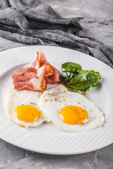 Breakfast, fried eggs, bacon, prosciutto, fresh salad on plate on grey table surface. Healthy food