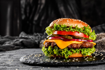 Beef burger with cheese, tomatoes, red onions, cucumber and lettuce on black slate over dark background. Unhealthy food - obrazy, fototapety, plakaty