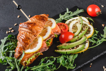 Croissant sandwich with red fish, avocado, fresh vegetables and arugula on black shale board over black stone background. Healthy food concept