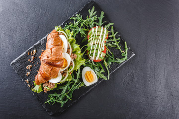 Croissant sandwich with tuna, avocado, fresh arugula and greens on black shale board over black stone background. Healthy food concept