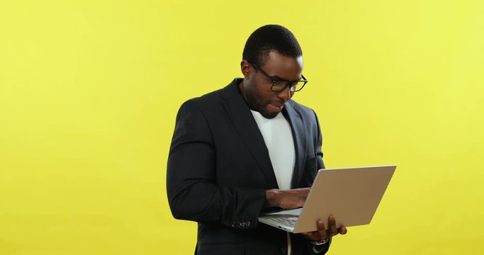 African American young businessman in glasses standing on the yellow background with a laptop computer.
