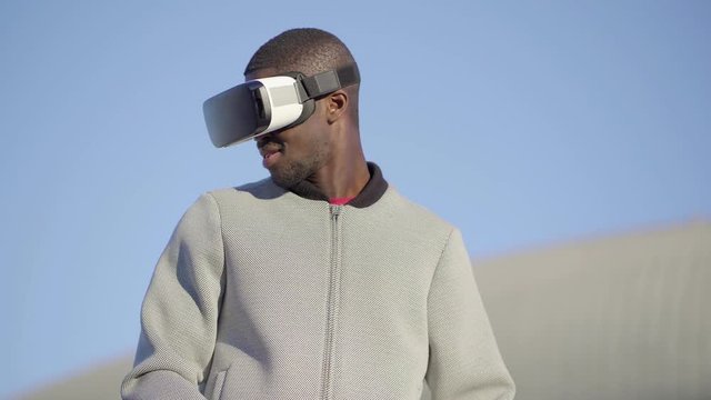 Young Afro-American short-cut muscular man in grey jacket wearing virtual reality glasses in park, managing new reality with hands, turning around. Dolly shot. High technology, VR concept