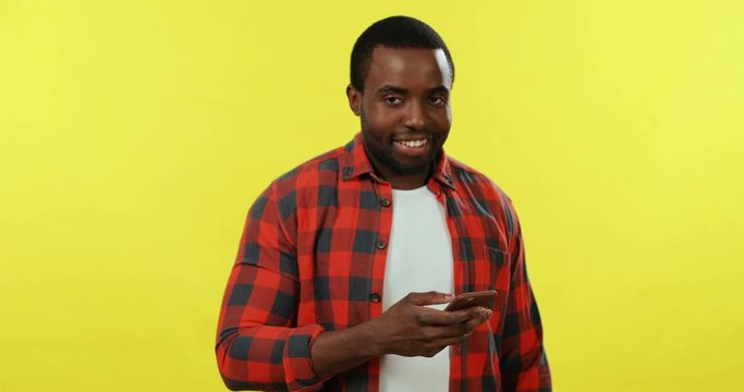 Portrait of the young cheerful African American man texting a message on the smartphone and smiling to the camera on the yellow background.