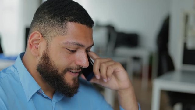Close up shot of handsome young man talking on phone. Smiling young bearded guy wearing blue shirt talking with friend through smartphone. Communication and technology concept