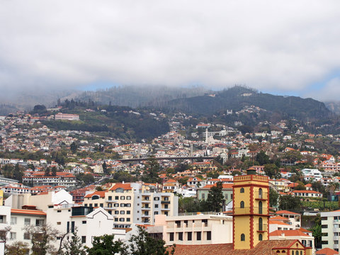 cityscape view of funchal in madeira with typical white painted buildings old church and bridge with tree covered mountains under white clouds