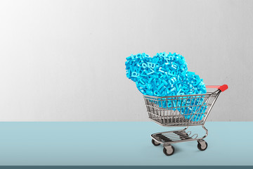 Big data and cloud computing concept. Shopping cart with cloud of blue letters and numbers on table and concrete background, side view.
