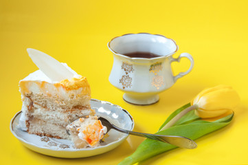 A cup of tea, a plate with a piece of cake, a teaspoon and a tulip flower on a yellow background
