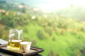 Chinese tea in glass tea pot and painted ceramic cup on wooden tray at black terrace in front of blurred mountain, green forest, white clouds and sky with copy space.