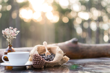 Selective focus of black coffee in white ceramic coffee cup with coffee beans in the sack, pine cones and flowers on wooden table with blurred bokeh forest background