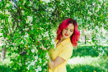 Beautiful and young woman with bright red hair and ref lips standing next to a blooming apple tree...
