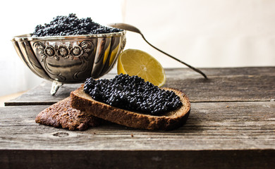 black caviar on rye bread with a lemon and a silver spoon on a background of moss and wood.