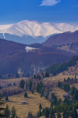 Rural scene in Romania with small houses placed on hills in the foreground between trees and a mountain peak in the background covered in snow