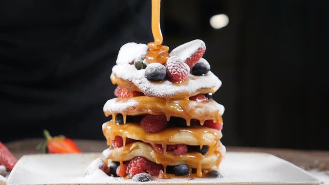 Cooking art. Chef spreading caramel on top of stack of fresh fluffy pancakes decorated with forest berries. Homemade Healthy Vegan Fluffy American Style Pancakes with Fresh Fruits. Pouring honey on