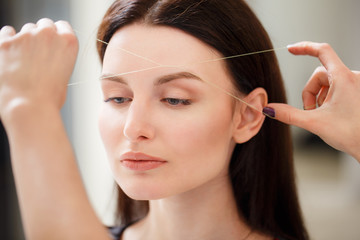 Cosmetologist plucks client eyebrows by thread. Close up