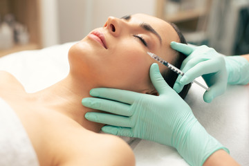 Woman lying with closed eyes and getting vitamin injection into the face skin