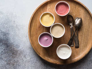 Fresh berries ice cream of different colors in paper cups and metal spoons on round wood tray on a gray background. Top view. copy space