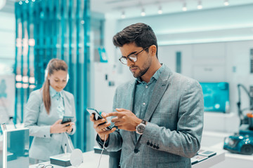 Serious mixed race unshaven man in formal wear and with eyeglasses trying out smart phone he want to buy. In background woman choosing smart phone.