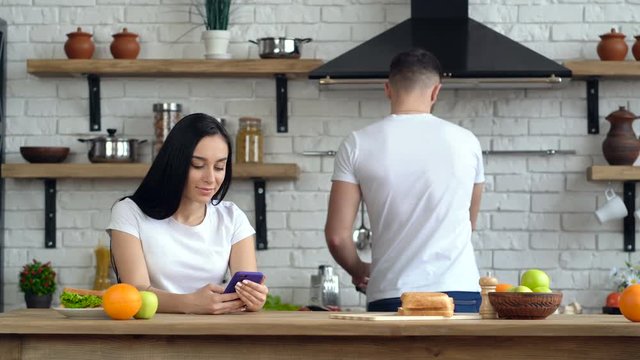 Young couple at the kitchen. Attractive woman holding and looking at the smartphone. Young man preparing coffee and giving to her.