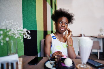 Сheerful african american young woman in summer dress at cafe with milkshake and dessert.