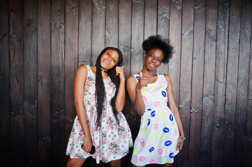 Fototapeta na wymiar Two black african girlfriends at summer dresses posed against dark wooden background and showing thumb up fingers.