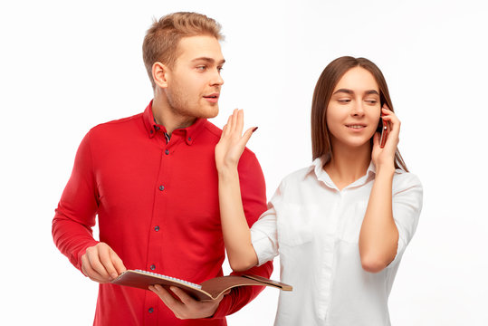 a young brunette girl in a white blouse is talking on the phone and makes a sign not to distract her young man in a red shirt with a document folder in his hands