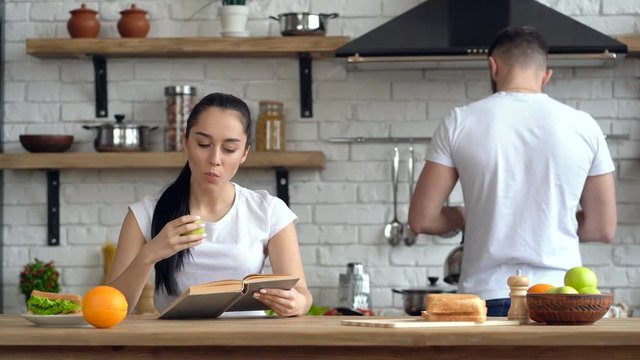 Couple at the kitchen. Breakfast, morning time. Young woman reading book and eating apple. Guy giving cup of coffee to her.