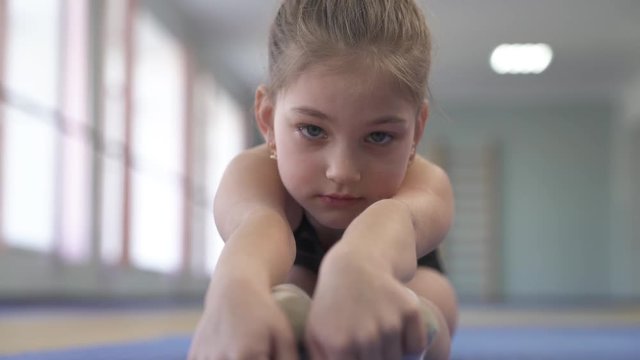 A close-up view of a little female gymnast doing stretching exercises (sitting in a pike position and stretching forward).