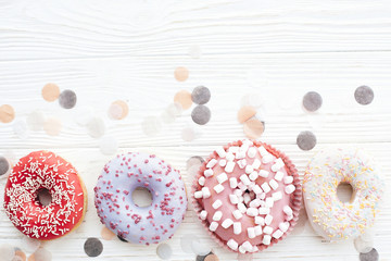 Delicious colorful donuts with sprinkles and marshmallows on stylish white table with confetti, flat lay, copy space. Party concept. No diet. Candy bar at wedding reception. Purple, pink donuts