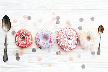 Fototapeta na wymiar Delicious colorful donuts with sprinkles and marshmallows on stylish white table with confetti and vintage spoon, flat lay. Party concept. No diet. Candy bar at wedding reception.