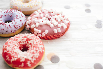 Delicious colorful donuts with sprinkles and marshmallows on stylish white table with confetti, copy space. Party concept. No diet. Candy bar at wedding reception. Purple, pink donuts