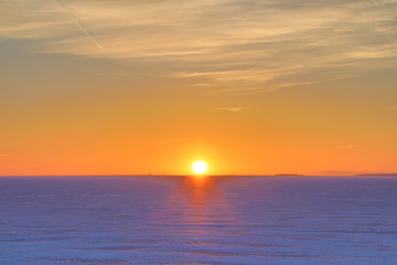 The sunset or sunrise. The cloudy sky cloured in red, orange, crimson, purple, violet and blue bright and vivid coloures with setting or rising sun over the sea, lake or bay covered with ice and snow