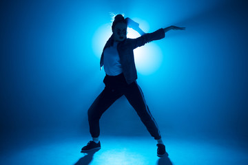 Young woman dressed in street fashion wear dancing hip-hop style over studio blue light background with flare effects