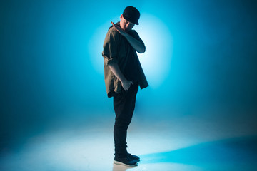 Sporty modern style hip-hop dancer dressed in urban style wear shows his dance on blue studio...